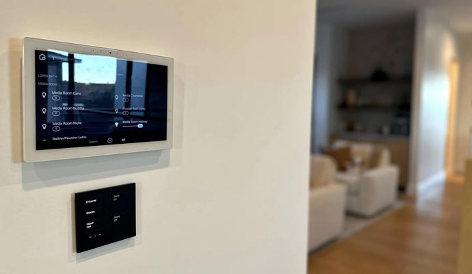 Wall-mounted smart home control panel displaying media room settings with a secondary switch panel below in a modern living space