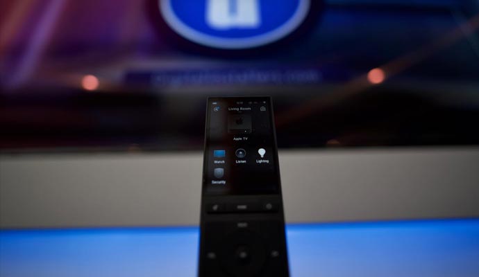 Close-up of a smart remote control with options for lighting and security in front of a large screen display.